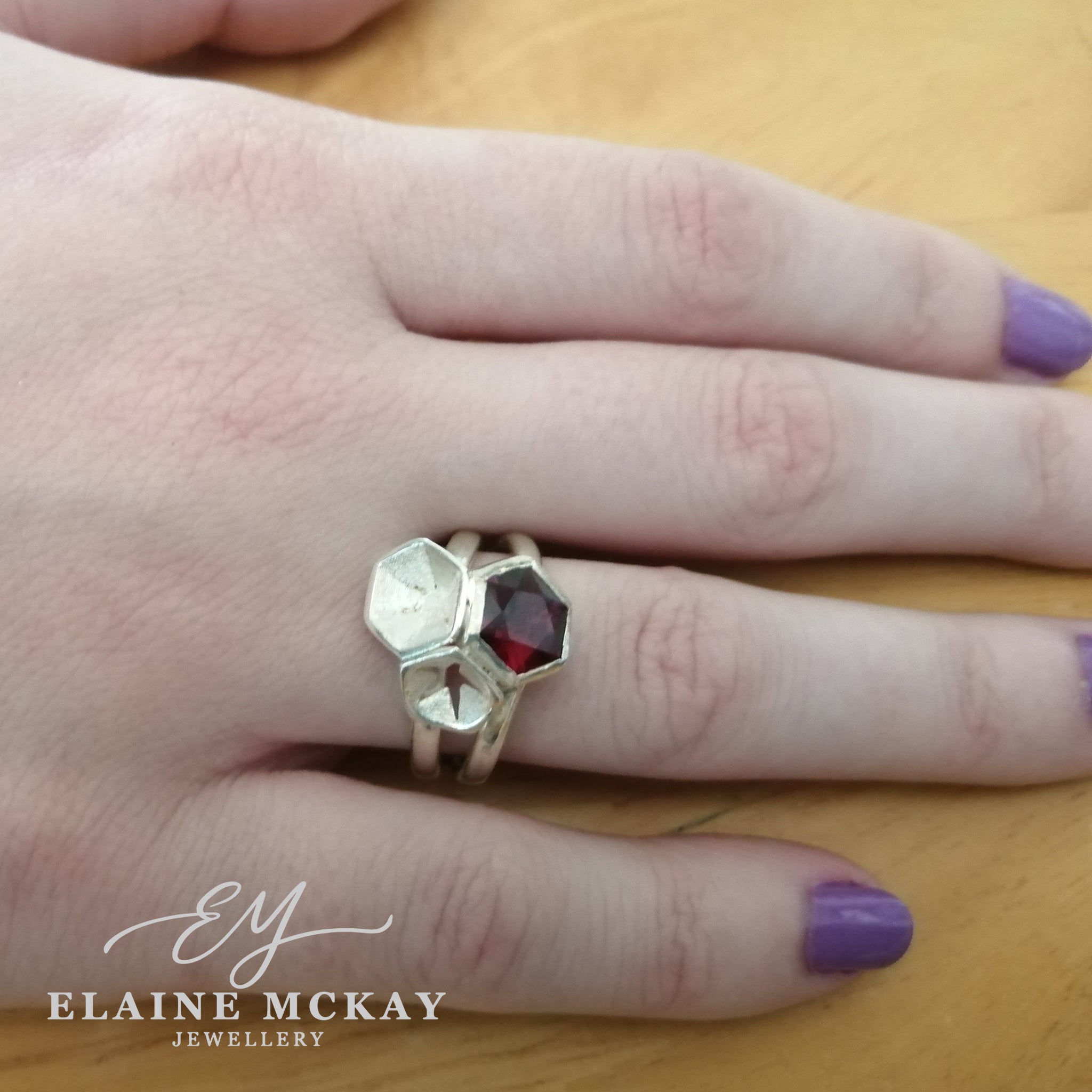 Hand wearing a silver ring with a red garnet gemstone on a wooden surface, branded 'Elaine McKay Jewellery'.